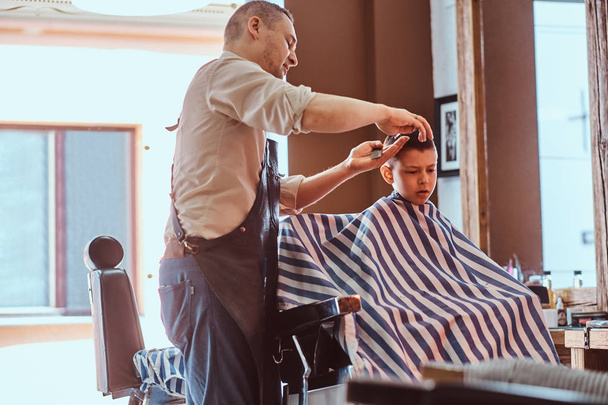 Pequeño chico encantador está consiguiendo corte de pelo de moda de peluquero expirienced en salón de peluquería de moda
 - Foto, imagen