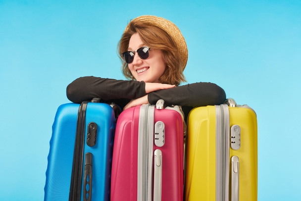 happy girl in sunglasses and straw hat near multicolored travel bags isolated on blue - Foto, Imagem