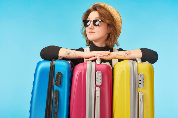smiling girl in sunglasses and straw hat looking away near multicolored travel bags isolated on blue - Foto, immagini