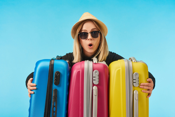 shocked girl in sunglasses and straw hat near multicolored travel bags isolated on blue - Foto, Imagem