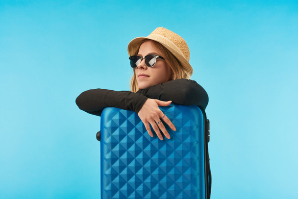chica rubia soñadora en gafas de sol y sombrero de paja cerca de la bolsa de viaje azul aislado en azul
 - Foto, Imagen