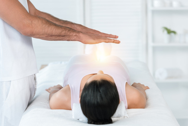 bijgesneden weergave van genezer zetten handen boven lichaam van brunette vrouw liggend op massagetafel - Foto, afbeelding