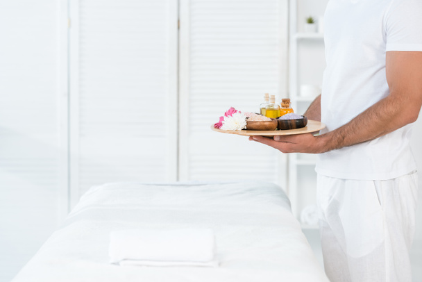 cropped view of man holding tray with bottles of oil, bowls with sea salt and flowers in spa center  - Foto, Imagem