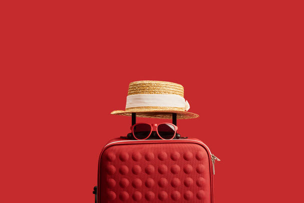 red travel bag with straw hat and sunglasses isolated on red  - Фото, зображення
