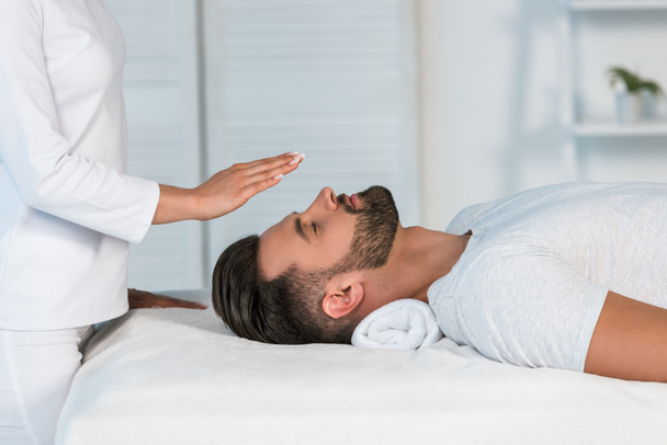 visão cortada de curandeiro colocando as mãos acima da cabeça de homem bonito na mesa de massagem
 - Foto, Imagem