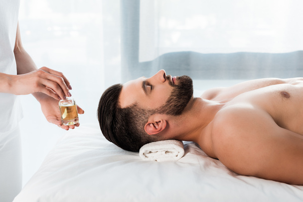 обрезанный вид массажиста, держащего бутылку с маслом рядом с красивым мужчиной с закрытыми глазами
 - Фото, изображение