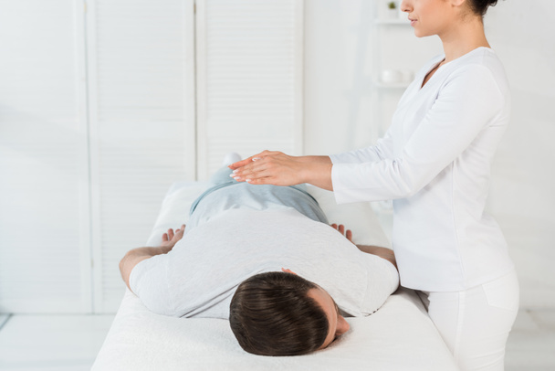 visão cortada de curandeiro colocando as mãos acima das costas do homem na mesa de massagem
 - Foto, Imagem