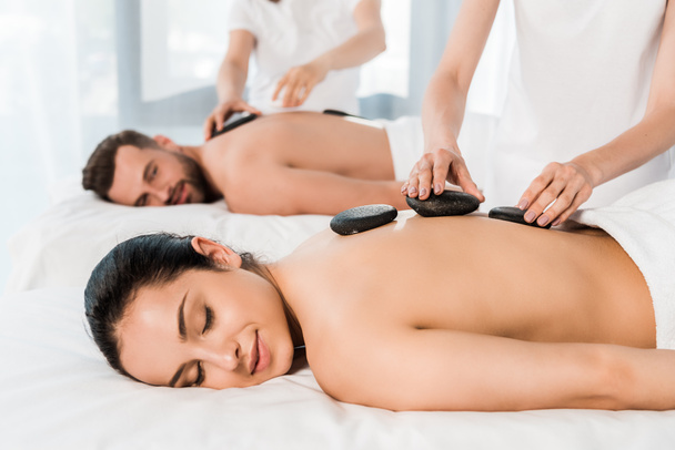 selectieve focus van masseur het doen van hete stenen massage naar aantrekkelijke vrouw met gesloten ogen in de buurt van bebaarde man  - Foto, afbeelding