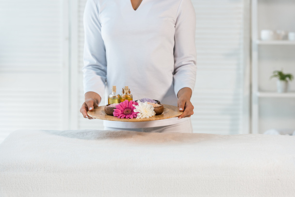 cropped view of young masseur holding tray with bottles of oil, sea salt and flowers - Foto, Bild