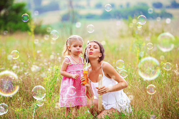Mother and little girl daughter child blowing soap bubbles outdoor. Parent and kid having fun on meadow. Happy and carefree childhood. Good family relations - Φωτογραφία, εικόνα