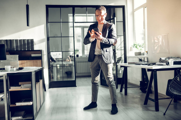 Liikemies seisoo toimistossa ja käyttää älypuhelintaan
 - Valokuva, kuva