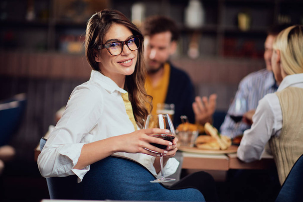 Portrait of gorgeous brunette looking at camera and holding glass with red wine while sitting at restaurat. In background friends eating diner. - 写真・画像