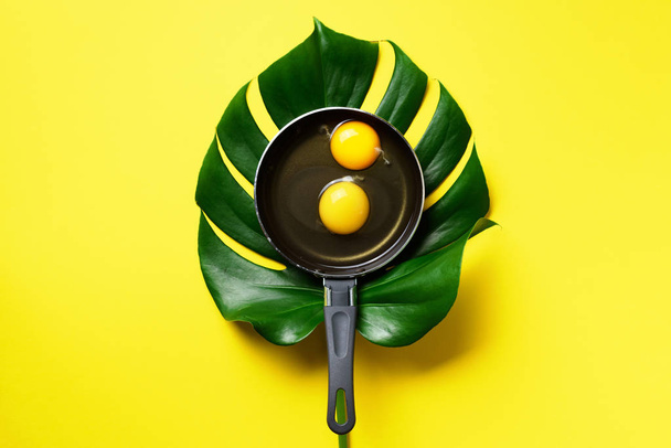 Organic egg in pan over monstera leaf on yellow background. Minimalistic food concept. Top view. Creative layout - Fotó, kép