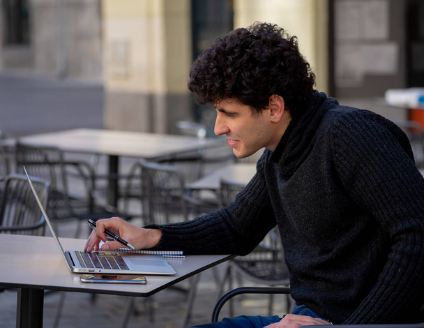 Красивый молодой человек, работающий за ноутбуком на террасе в кофейне европейского города. Студент чувствует себя счастливым и довольным. В путешествиях по миру онлайн-образование и концепция технологий
. - Фото, изображение