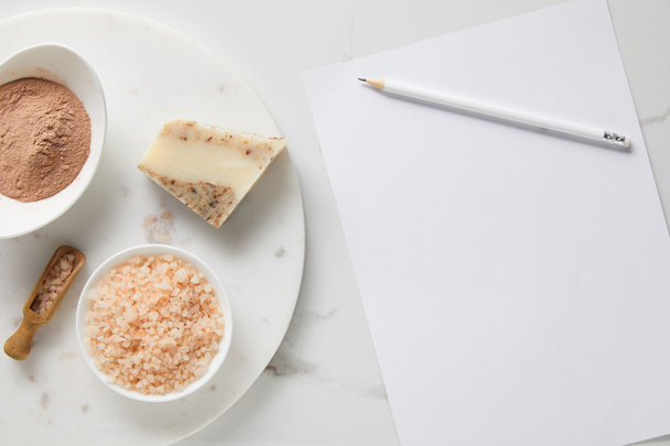 top view of clay powder, soap and sea salt on marble table near white paper and pencil - 写真・画像