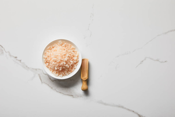top view of sea salt in bowl near wooden spatula on marble table - Foto, afbeelding