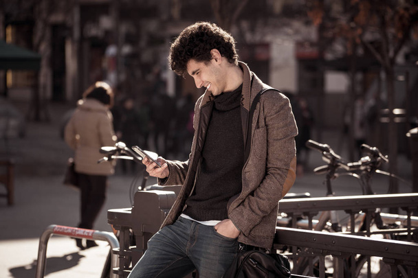 Fashion college man in his twenties happy checking blog or chatting on the internet with friend on smart mobile phone app in european city outdoors city. In communication technology and travel Europe. - Photo, Image