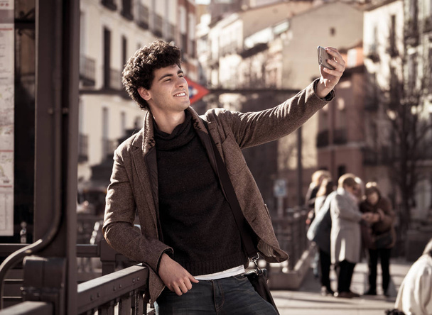 Excited trendy young man taking selfie or blogging on smart mobile phone on holidays in european city street or university campus. Holidays and technology Connection and social media concept. - Foto, imagen