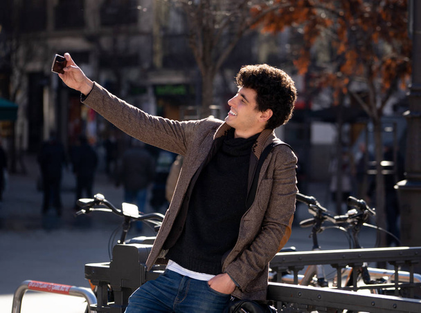 Excited trendy young man taking selfie or blogging on smart mobile phone on holidays in european city street or university campus. Holidays and technology Connection and social media concept. - Фото, зображення