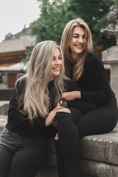 Two beautiful girls sitting at the fountain. Friends wearing in black clothes playing near fountain. Young tourists talking and laughing in the old city. Positive ladies enjoying summer vacations  - Foto, immagini