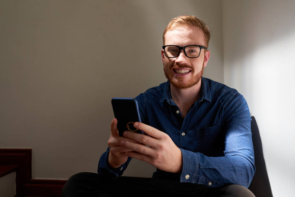 Portrait of handsome red haired man in eyeglasses sitting on stairs using mobile phone and smiling at camera - Foto, imagen
