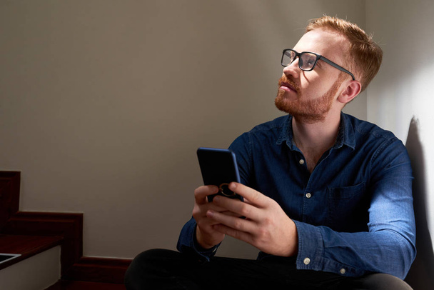 Vörös hajú férfi szemüvegek nyugszik lépcsőn, és keres átgondolt, miközben ő használ a mobiltelefonján - Fotó, kép