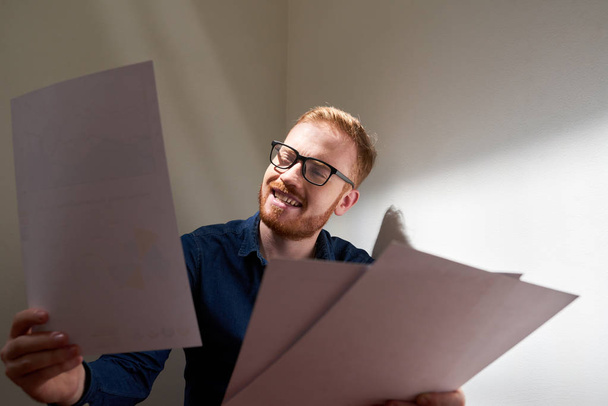 Red haired businessman wearing eyeglasses reading documents in his hands and smiling - Foto, Imagem
