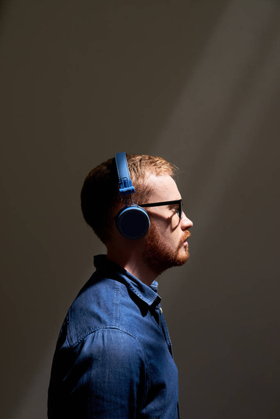 Zijaanzicht afbeelding van jonge roodharige man in draadloze koptelefoon en in brillen staande en luisteren naar muziek over grijze achtergrond - Foto, afbeelding