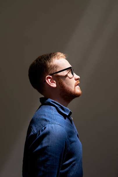 Side view image of young red haired businessman standing in eyeglasses and jeans jacket isolated on grey background - Foto, Imagen
