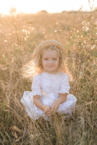 Cute little girl with blond  hair in a summer field at sunset with a white dress with a straw hat - Foto, Imagen