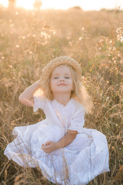 Cute little girl with blond  hair in a summer field at sunset with a white dress with a straw hat - Valokuva, kuva