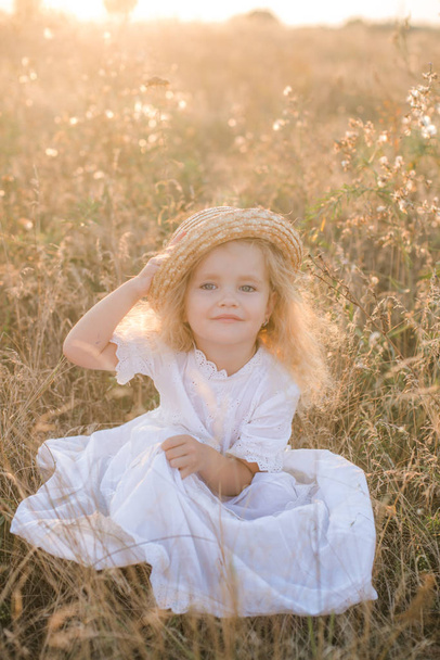 Cute little girl with blond  hair in a summer field at sunset with a white dress with a straw hat - Фото, изображение