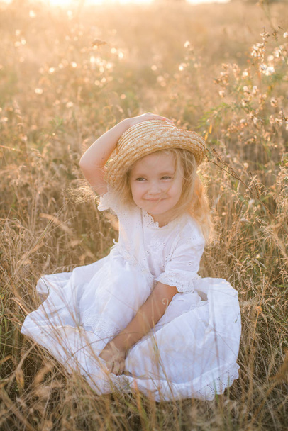 Cute little girl with blond  hair in a summer field at sunset with a white dress with a straw hat - Zdjęcie, obraz