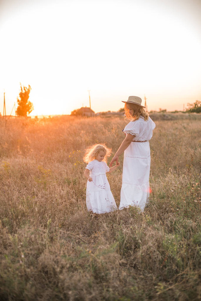 mother and daughter holding hands and walking by green meadow with sunset sky on background - Fotoğraf, Görsel