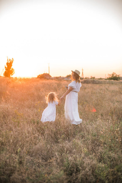 mother and daughter holding hands and walking by green meadow with sunset sky on background - 写真・画像