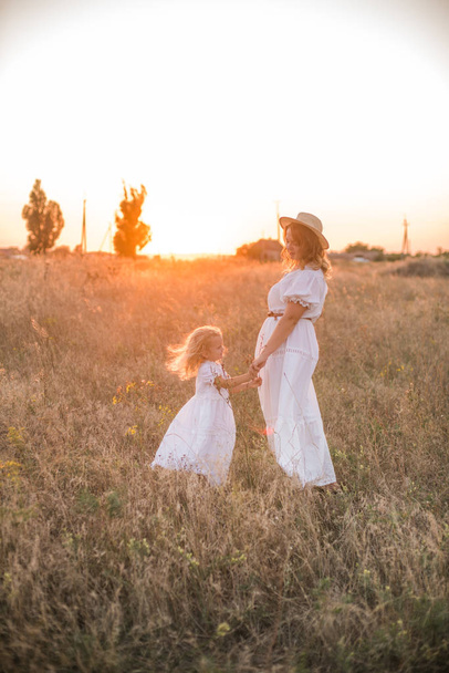 mother and daughter holding hands and walking by green meadow with sunset sky on background - Foto, Bild