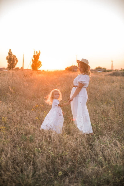 mother and daughter holding hands and walking by green meadow with sunset sky on background - Valokuva, kuva