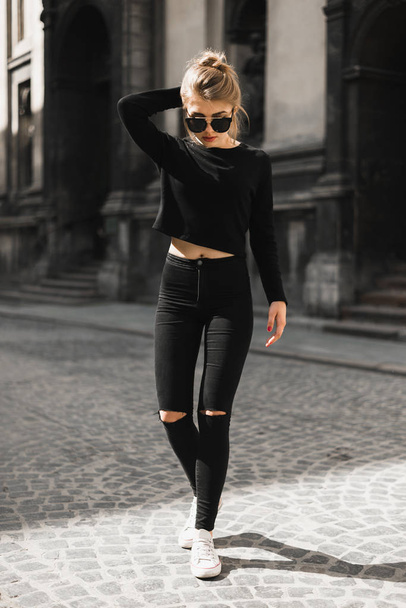 Outdoor portrait of a pretty woman walking near old buildings on a sunny day. Young female in black clothes strolls across street. Trendy girl enjoying summertime in the quaint old city - Photo, image
