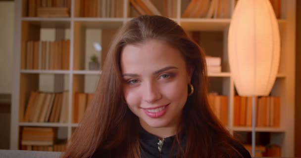 Close-up portrait of young female student smiles into camera seductively on bookshelves background. - Filmagem, Vídeo
