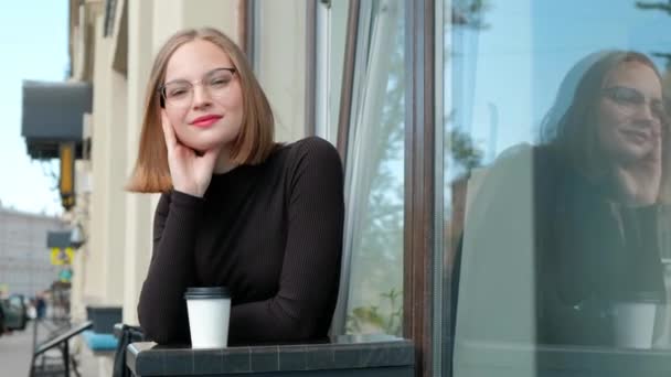 Attractive red-haired young woman with short haircut, a four-sided, with glasses and brown turtleneck, is sitting on bench at table in street cafe. - Imágenes, Vídeo