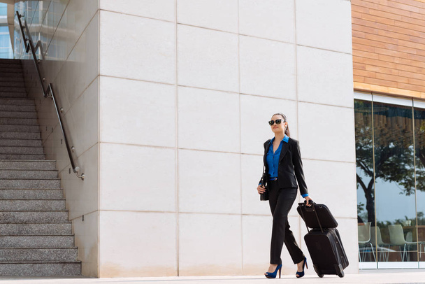 Деловая женщина в очках и костюме гуляет по улице с чемоданом
 - Фото, изображение