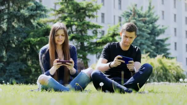 Happy couple play mobile game on smartphone, sitting on the grass in the park. Man win the game, woman lost. - Footage, Video