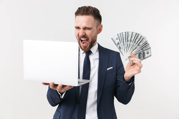 Emocionado hombre de negocios posando aislado sobre fondo de pared blanca sosteniendo ordenador portátil y dinero
. - Foto, imagen