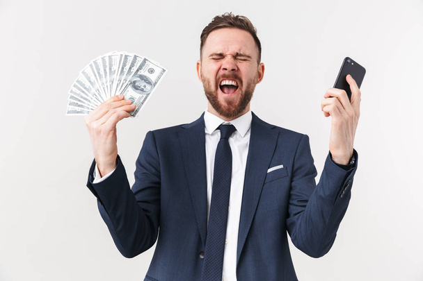 Excited businessman posing isolated over white wall background holding mobile phone and money. - Foto, imagen