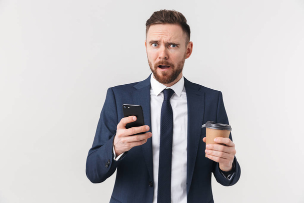 Displeased businessman posing isolated over white wall background drinking coffee using mobile phone. - 写真・画像