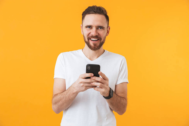 Ευτυχισμένος νέος όμορφος άντρας που παρουσιάζουν απομονωμένα πάνω από κίτρινο φόντο τοίχου χρησιμοποιώντας το κινητό τηλέφωνο. - Φωτογραφία, εικόνα