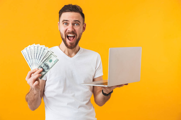 Happy young handsome man posing isolated over yellow wall background using laptop computer holding money. - Foto, immagini
