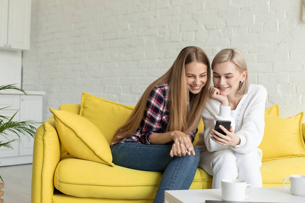 Two young attractive woman in casual clothes sitting on yellow sofa at home - Photo, Image