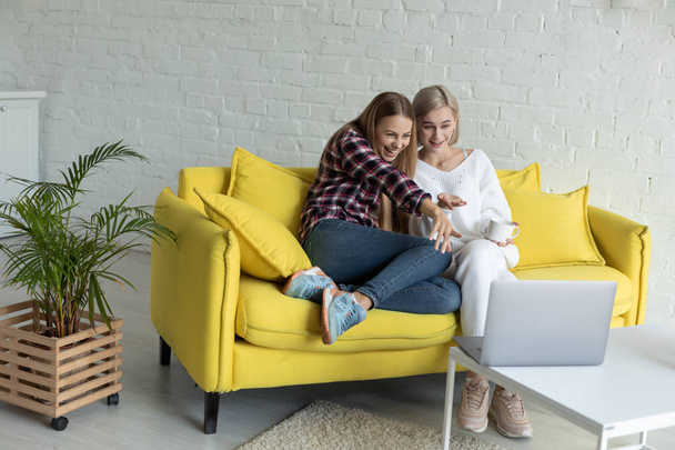 Happy young female lesbian couple in casual clothes sitting together on yellow sofa at home, are watching funny movie on laptop - Фото, изображение