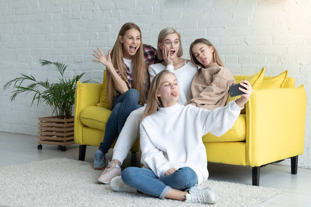 Happy atrakcyjna rodzina lesbijek w casual ubrania co selfie siedząc na żółtej kanapie w domu, blond córtet siedzi na podłodze z skrzyżowanymi nogami i trzymając telefon w ręku - Zdjęcie, obraz